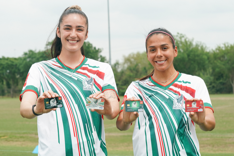 Imagen de dos jugadoras de la Selección Nacional de México llevan puesta la camiseta con la marca de Wells Fargo con el escudo de la Selección Nacional de México mientras sostienen cada una dos tarjetas frente a ellas.