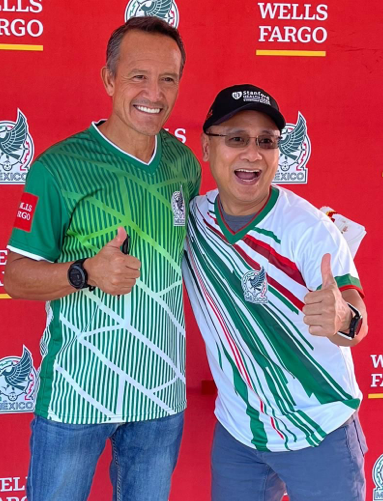 Imagen de un aficionado que se esta tomando una foto con un exjugador de la Selección Nacional de México, ambos parados frente a un fondo rojo de Wells Fargo.