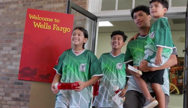 Foto de cuatro jóvenes parados frente a una sucursal de Wells Fargo con la camiseta verde de Wells Fargo que tiene el escudo de la Selección Nacional de México.
                  